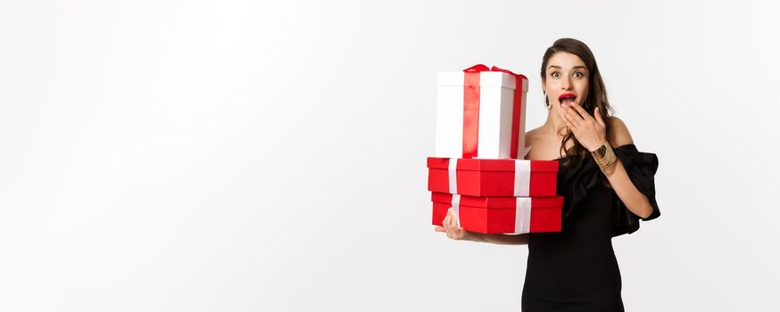 Celebration and christmas holidays concept. Woman holding xmas gifts and looking surprised, receive presents, standing over white background.