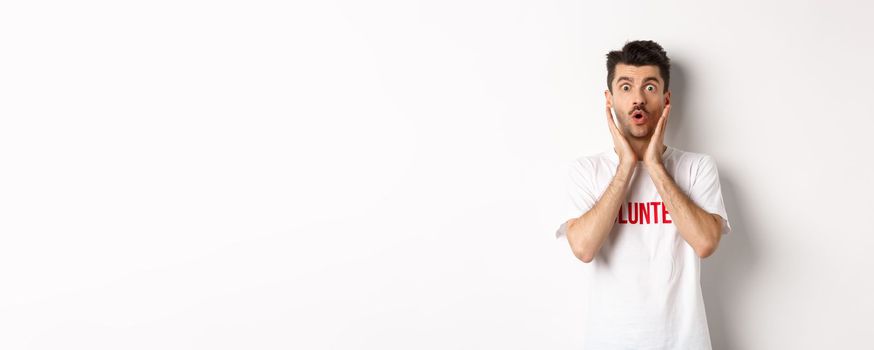 Shocked man in volunteer t-shirt gasping, staring at camera in awe, standing over white background.