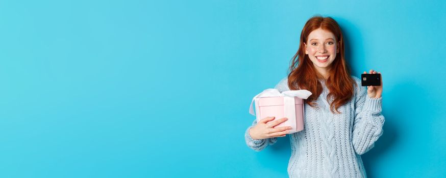 Happy redhead girl buying gifts with credit card, holding box with present and smiling, standing over blue background.