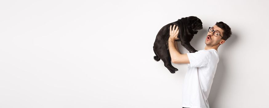 Image of handsome young man loving his pug. Dog owner holding puppy and smiling happy at camera, standing over white background.