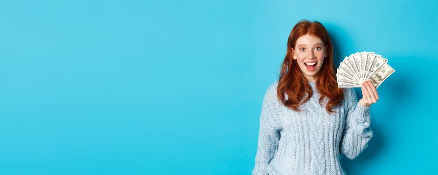 Happy redhead girl in sweater, staring excited at camera, showing money dollars, standing against blue background.