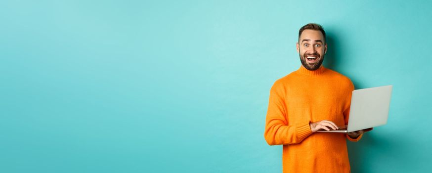 Excited smiling man working on laptop, staring at camera happy, standing in orange sweater against turquoise background.