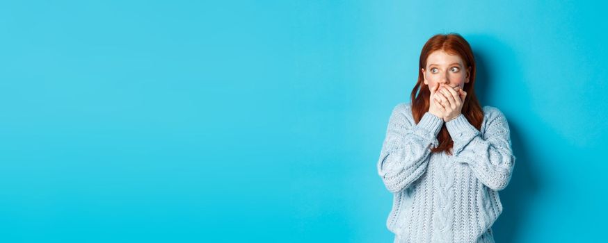 Shocked and anxious redhead girl staring left scared, covering mouth and gasping, standing over blue background in sweater.