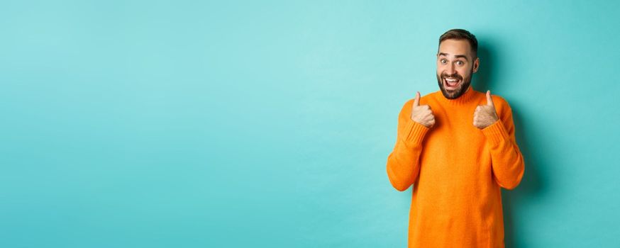 Excited young man with beard, showing thumbs up in approval, praise or recommend, standing over light blue background.