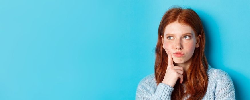 Headshot of troubled teenage girl thinking, looking bothered and frowning, standing against blue background.