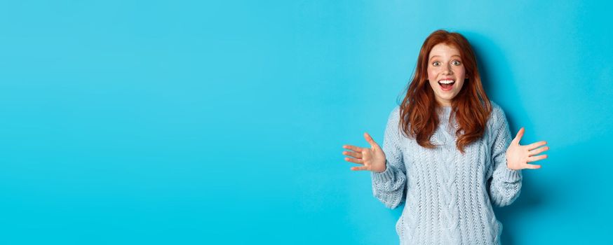 Excited redhead woman shaking hands, explaining big news, staring at camera amazed, standing against blue background.