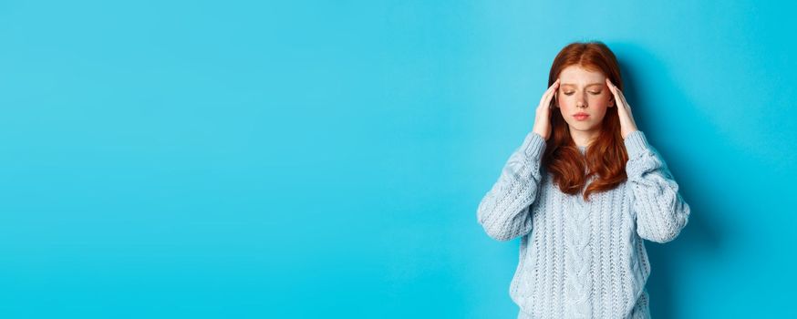 Distressed teenage redhead girl touching head, looking down with troubled face expression, standing against blue background, have problem.