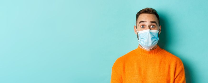 Covid-19, social distancing and quarantine concept. Close-up of young man in face mask staring at camera with surprised face, standing over turquoise background.