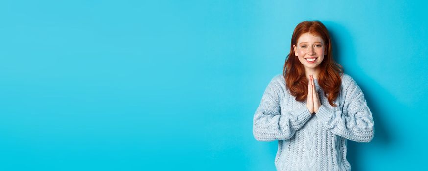 Cute redhead girl saying thank you, smiling and looking at camera, expressing gratitude, standing against blue background.