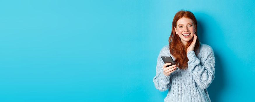 Beautiful redhead girl in sweater, smiling at camera, using mobile phone app, standing with smartphone against blue background.