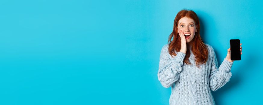 Amazed redhead girl looking at camera, showing smartphone screen, demonstrating mobile application, standing over blue background.