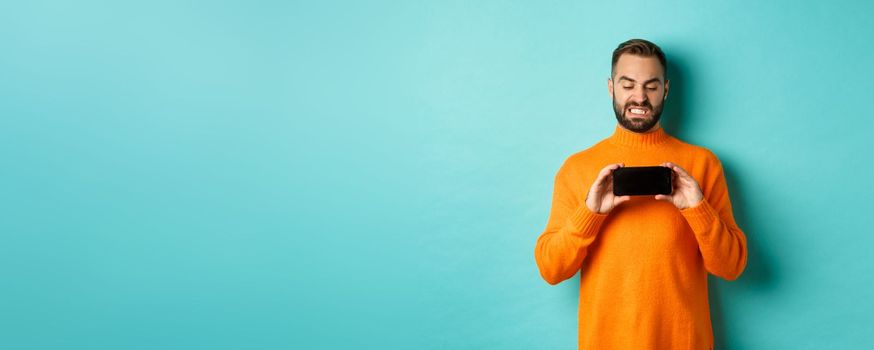 Image of man cringe at something on mobile screen, staring disgusted at display, showing smartphone, standing over light blue background.