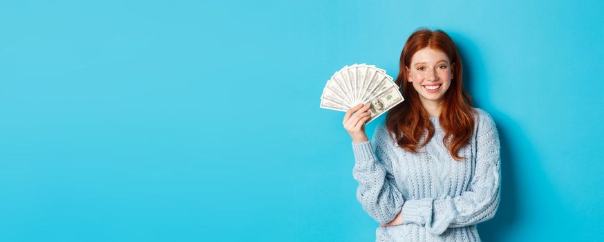 Happy and unbothered redhead woman holding money and smiling, looking confident and carefree, standing in sweater against blue background.