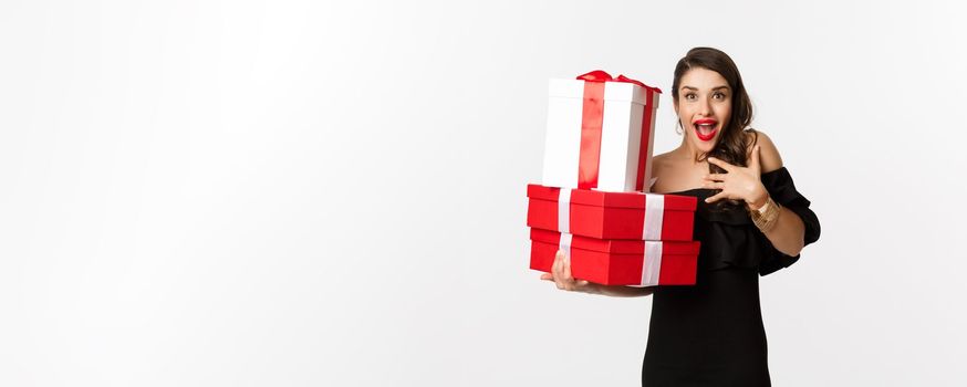 Celebration and christmas holidays concept. Excited and happy woman receive gifts, holding xmas presents and rejoicing, standing in black dress over white background.