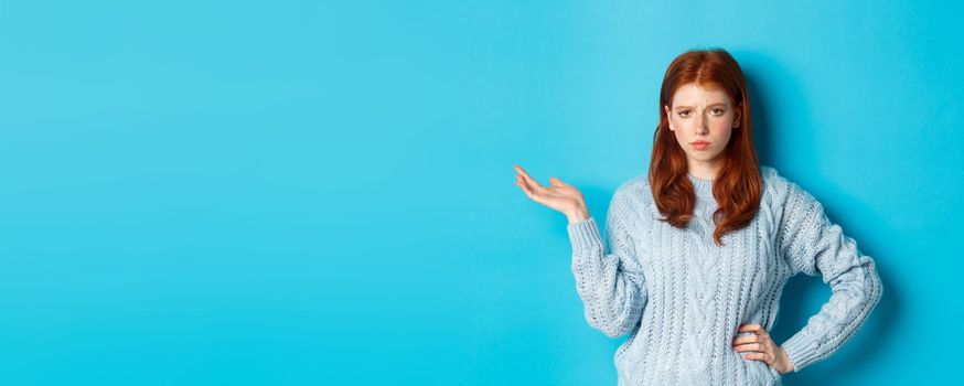 Skeptical teenage girl looking unamused, raising hand in so what gesture, staring at something with careless face, standing over blue background.