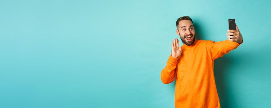 Happy young man video calling, talking online with mobile phone, saying hello to smartphone camera and waving hand friendly, standing over light blue background.