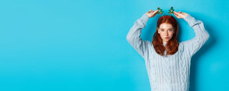 Winter holidays and Christmas sales concept. Beautiful redhead female model celebrating New Year, wearing funny party headband and sweater, smiling at camera.