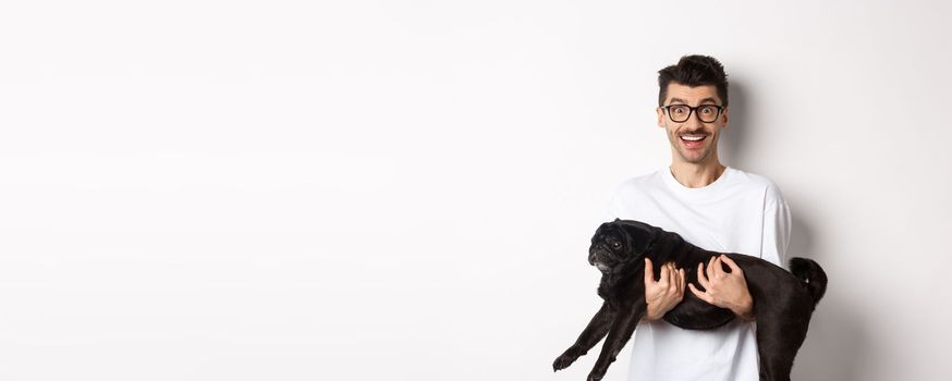 Happy young man in glasses holding black pug pet and staring at camera amused, smiling cheerful, standing over white background.