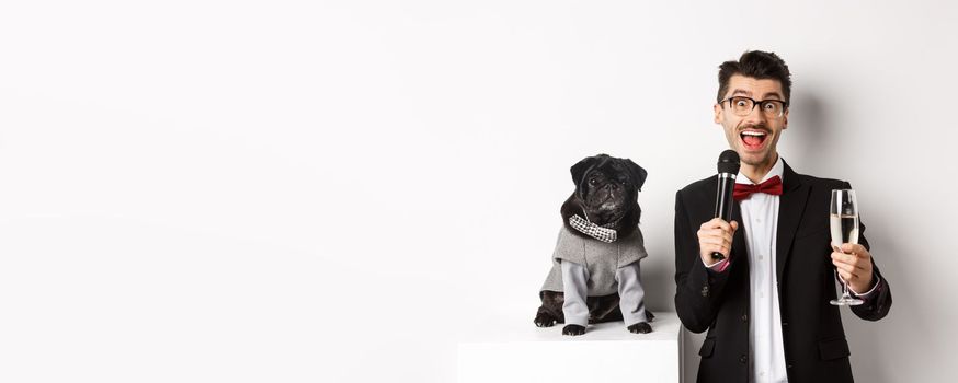 Happy young man giving a speech and raising glass champagne, standing with cute black dog in party costume over white background.