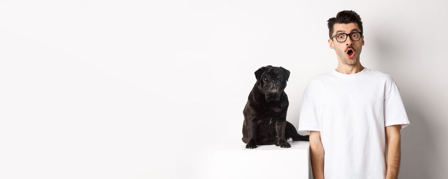 Image of dog owner and cute black pug looking at camera surprised and amazed, standing over white background.