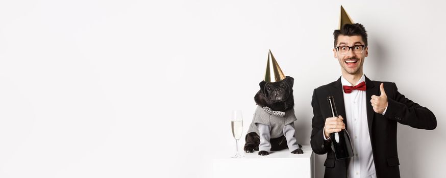 Small black dog wearing party hat and standing near happy man celebrating holiday, owner showing thumb-up and holding champagne bottle, white background.