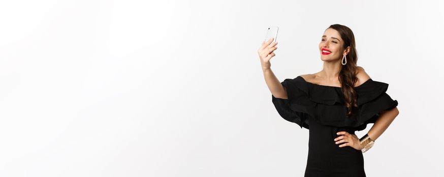 Beautiful woman in black dress taking selfie on party, standing over white background with smartphone.