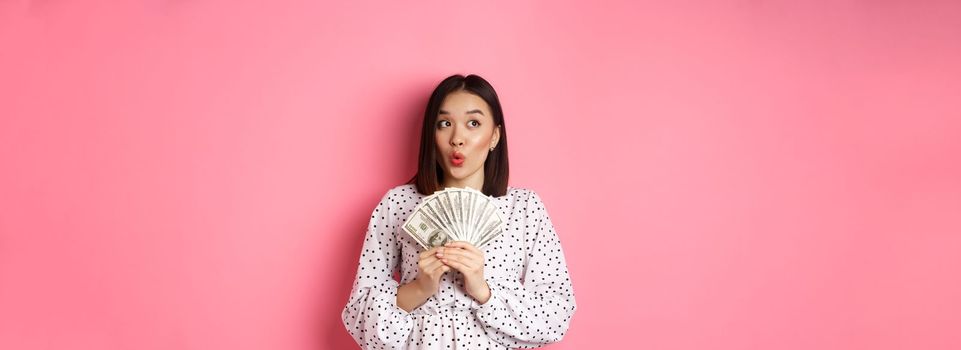 Shopping concept. Dreamy asian woman thinking, holding money dollars and looking aside thoughtful, standing over pink background.