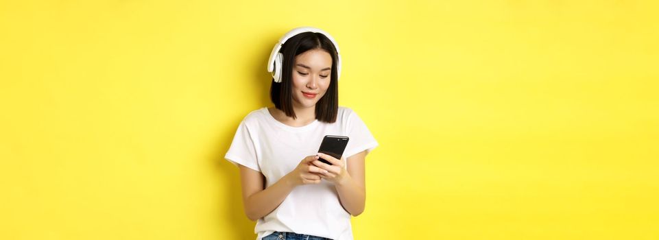 Modern asian girl listening music in wireless headphones, reading smartphone screen and smiling, standing in white t-shirt over yellow background.