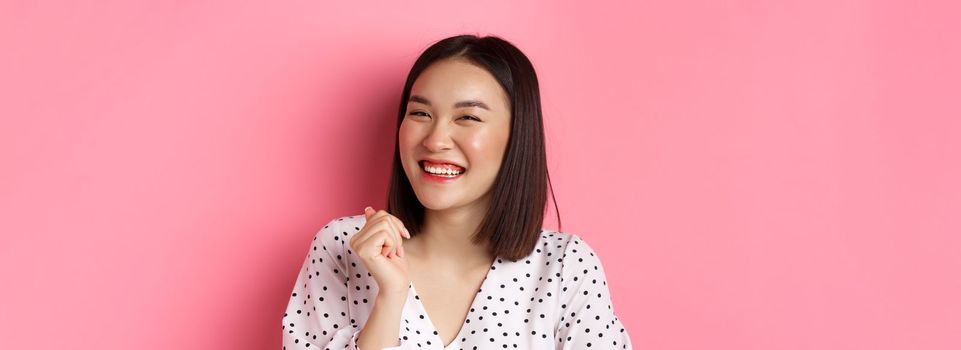 Beauty and lifestyle concept. Close-up of happy asian female laughing, looking happy and showing genuine emotions, standing over pink background.