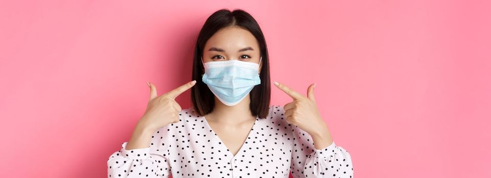 Covid-19, pandemic and lifestyle concept. Kawaii asian girl pointing fingers at her face mask, wearing preventive measures from coronavirus, smiling with eyes, pink background.