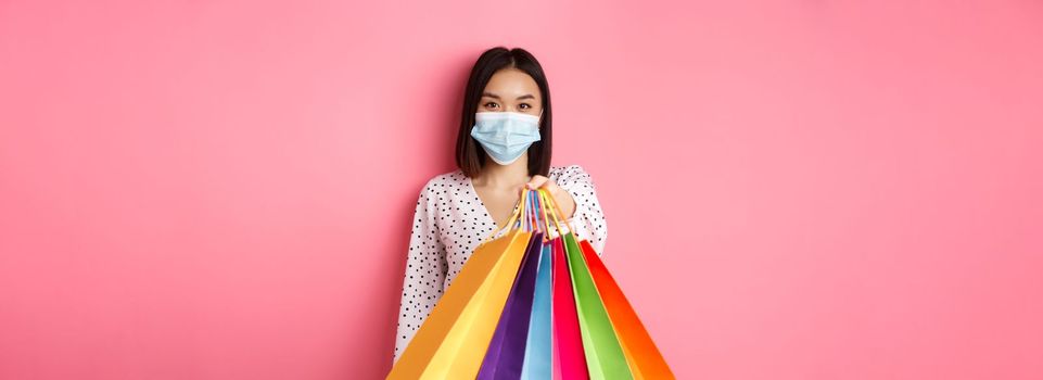 Covid-19, pandemic and lifestyle concept. Beautiful asian woman in face mask giving shopping bags, social distance in store, standing over pink background.