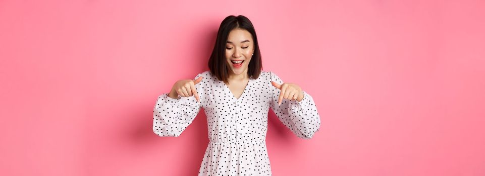 Romantic asian woman peeking down, pointing fingers at bottom, looking curious at product discount with happy smile, standing over pink background.