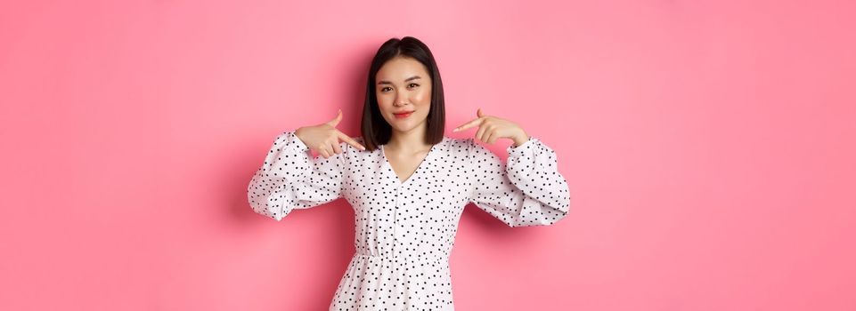Beautiful asian woman looking confident, pointing at herself and smiling, showing logo on chest, standing over pink background.