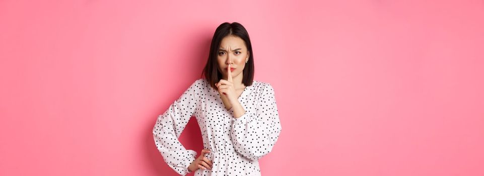 Angry asian female model shushing, frowning and hushing, showing taboo gesture, standing displeased against pink background, demand silence.