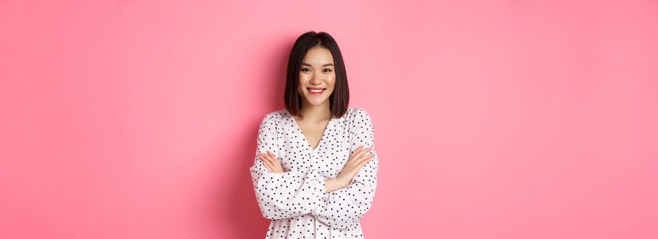 Pretty young asian woman in spring dress smiling, cross arms on chest and looking confident, standing over pink background.