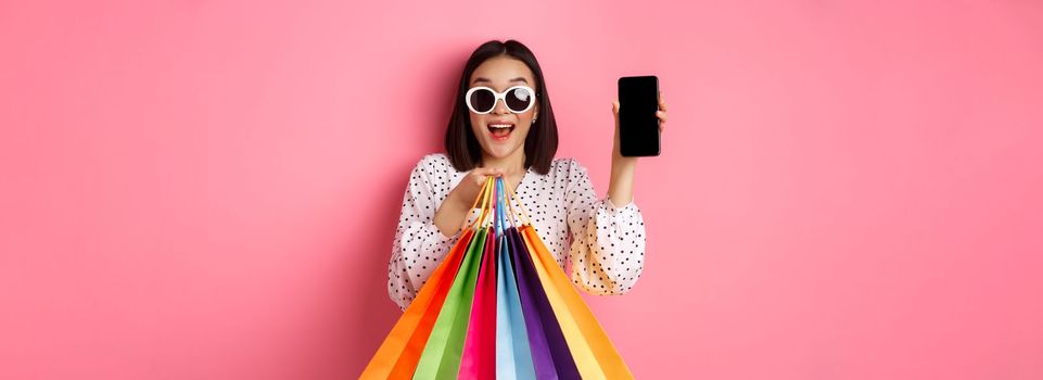 Attractive asian woman showing smartphone app and shopping bags, buying online via application, standing over pink background.