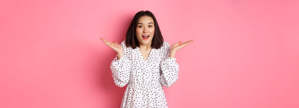 Image of surprised korean girl in dress, female model staring at camera and gasping amazd, standing over pink background.