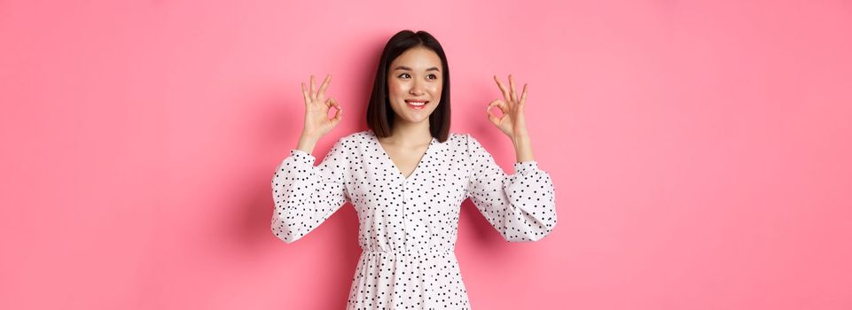 Pretty young asian woman in dress showing okay sign, praising and showing approval, looking satisfied, standing against pink background.