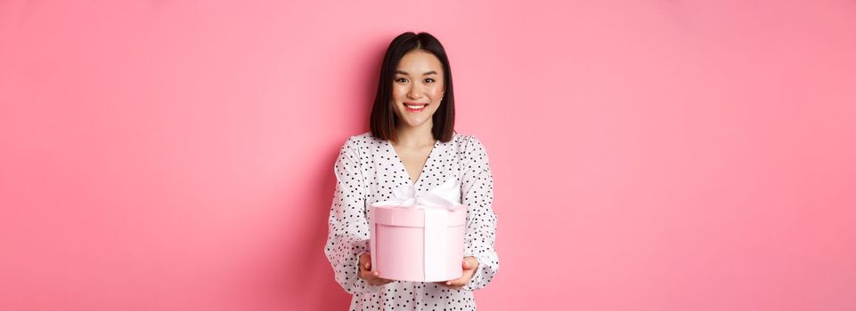 Beautiful asian woman wishing happy holidays, giving you gift in cute box, standing against pink background.