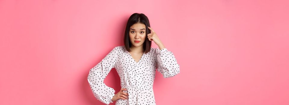 Image of attractive asian lady scolding person, pointing at head and staring at camera disappointed, are you stupid gesture, standing over pink background.