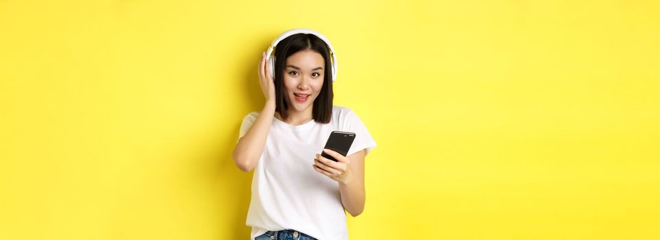 Modern asian woman listening music in wireless headphones, reading smartphone screen and smiling, standing in white t-shirt over yellow background.