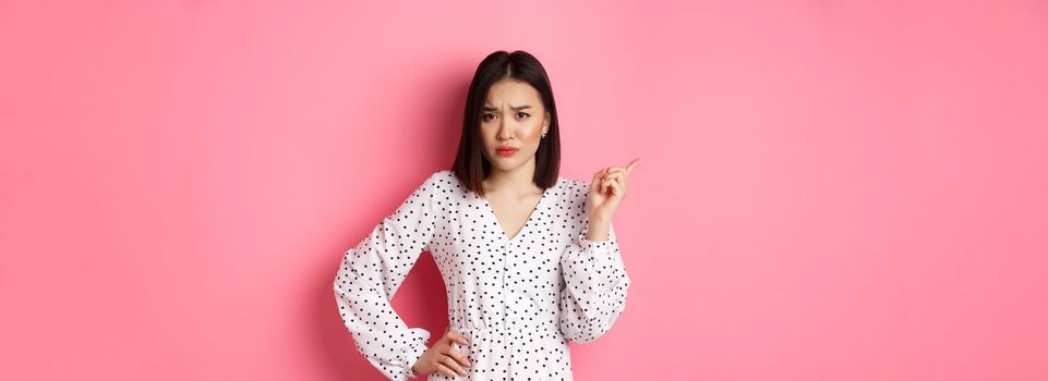 Disappointed asian girlfriend complaining, staring at camera and whining, pointing at upper left corner copy space, standing in dress over pink background.