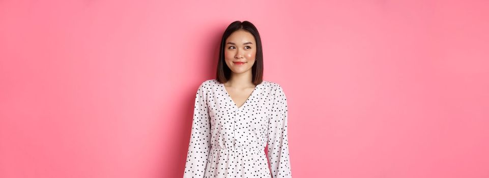 Cute asian woman smiling, looking left at copy space, standing on romantic pink background.