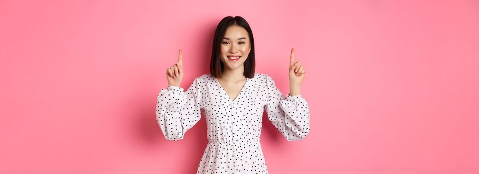 Beautiful Korean young woman in dress showing announcement, pointing fingers up at promo copy space, smiling happy, standing over pink background.