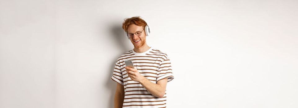 Technology concept. Young redhead hipster in glasses, listening music in headphoes, reading message on smartphone, white background.