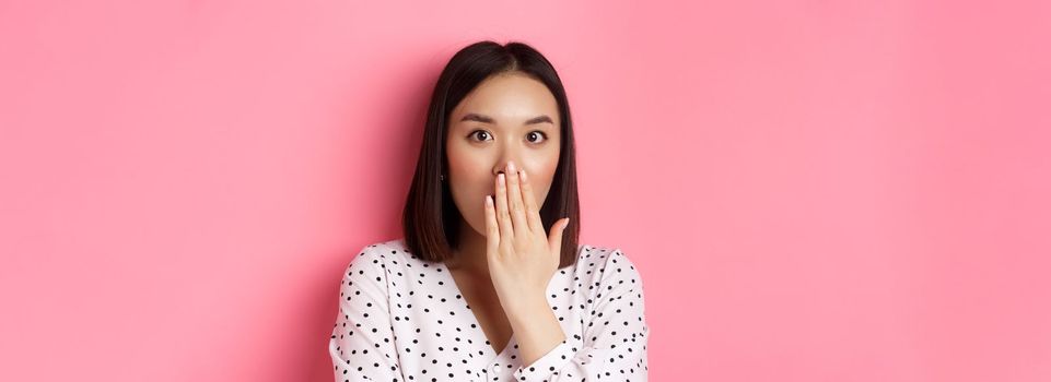 Image of shocked asian girl gossiping, gasping and cover mouth, stare at camera with complete disbelief, standing over pink background.