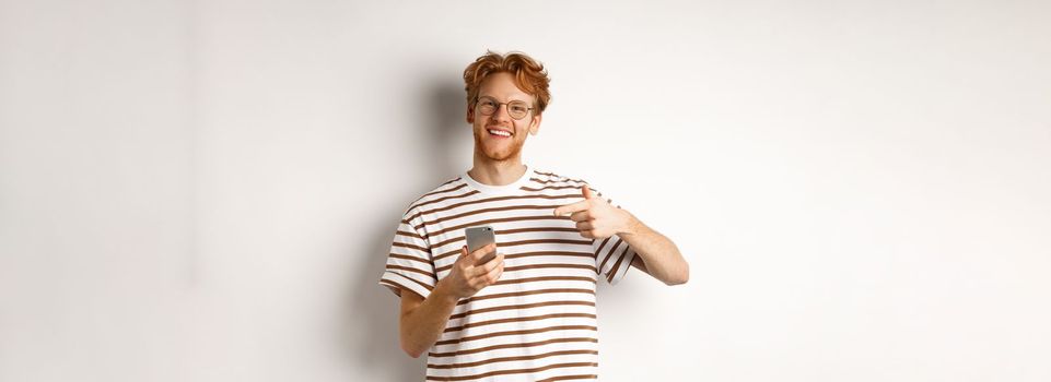 Technology and e-commerce concept. Young man with red messy hair, wearing glasses and t-shirt, pointing finger at smartphone and smiling satisfied, white background.