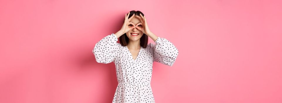 Carefree young asian woman looking through hand binoculars at camera, staring at discounts, standing over pink background.