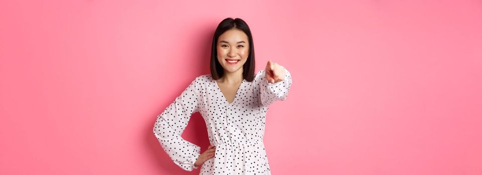 Confident asian woman in dress pointing at you, smiling and staring at camera, standing against pink background. Copy space