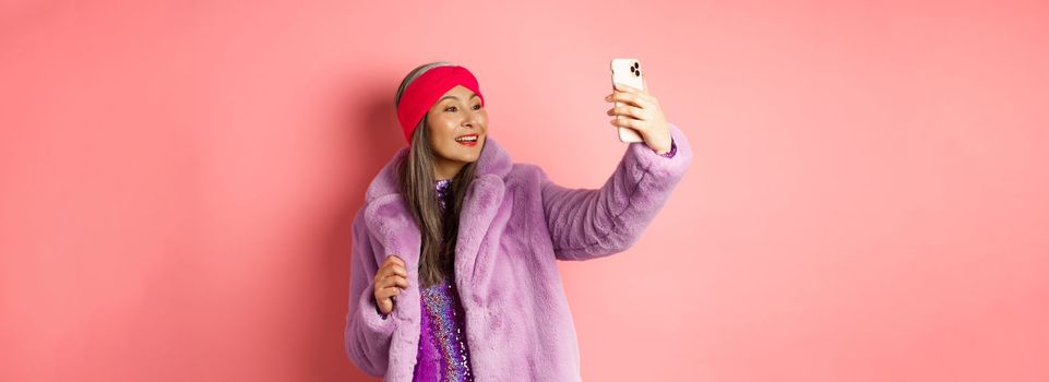 Fashion concept. Beautiful asian senior lady taking selfie on smartphone, posing in party glittering dress and purple faux coat, standing over pink background.